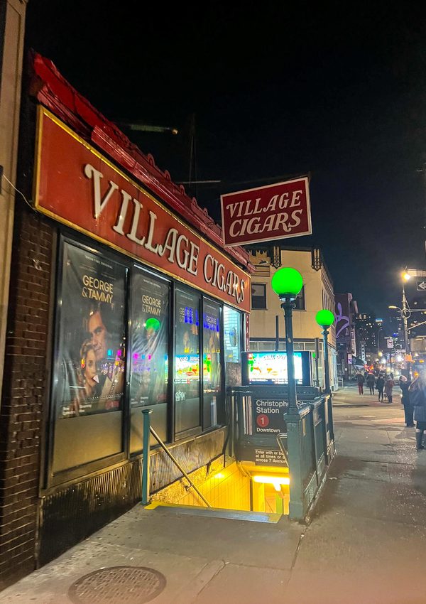 Village Cigars storefront at night, featuring the Christopher Street 1 station subway entrance stairs for navigating the New York City subway.