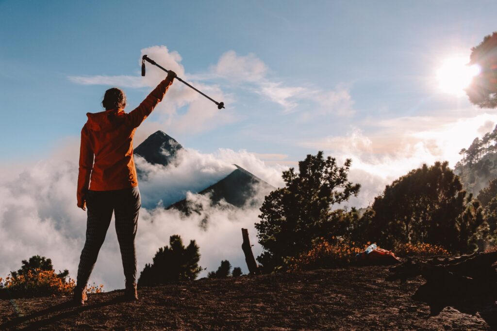 This is an image of a woman standing on a mountain top holding her walking stick as she looks at the mountains in the background surrounded by clouds.