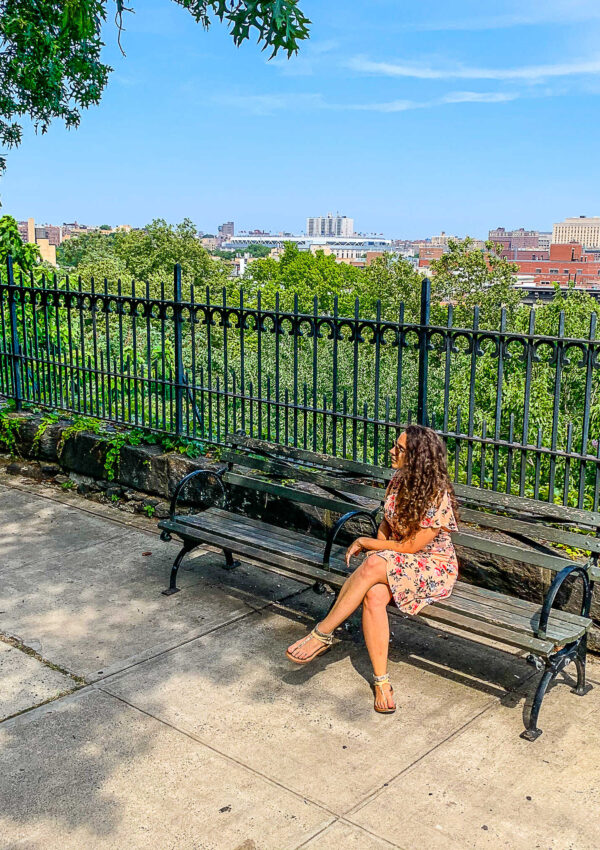 This is an image of a woman in a pink dress sitting on a bench and  looking away from the camera in Harlem, New York City. Yankee Stadium is behind her in the distance.