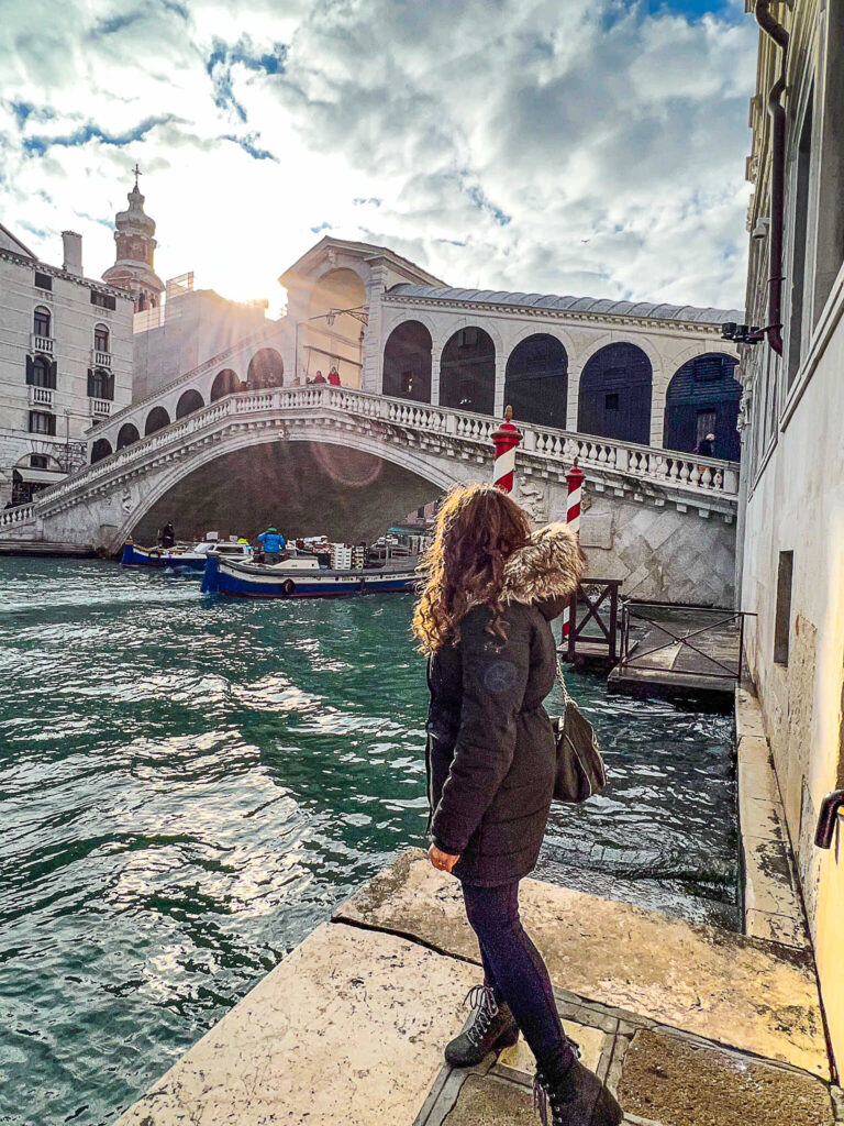 This is an image of a woman with her back to the camera looking at the Rialto Bridge in Venice, Italy. The sun is shining through the clouds casting a beautiful light.