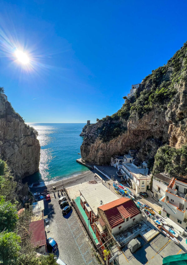 This is an image of a fiord on the Amalfi Coast. The sun is shining, there are cars and kayaks on shore next to a building, and the water is rolling in.
