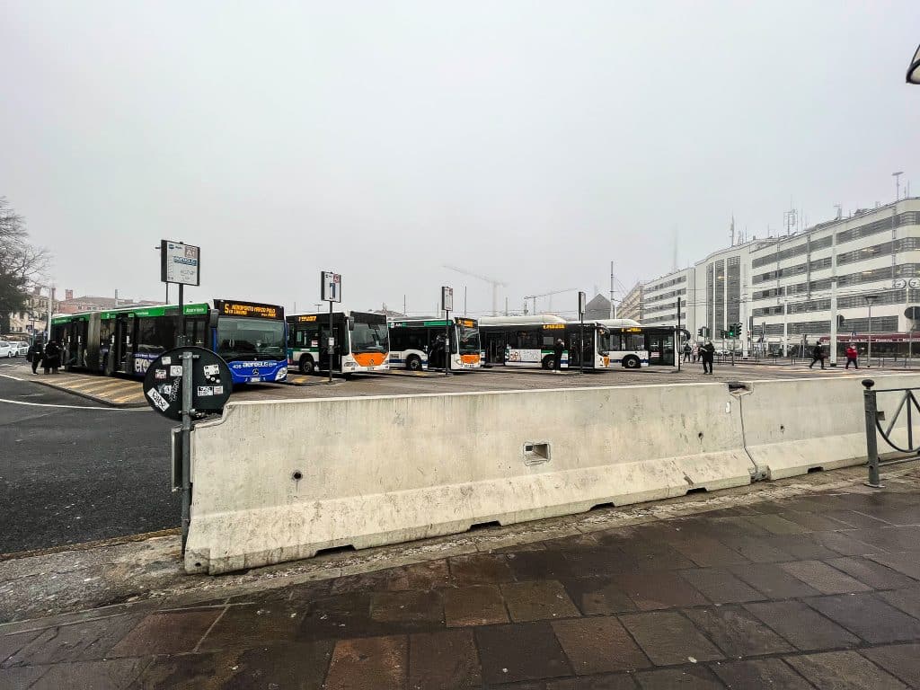 Several city buses are parked at a bus terminal on a cloudy day, with a concrete barrier and a parking structure visible in the background, prompting the question: are there cars in Venice unlike this busy terminal?.