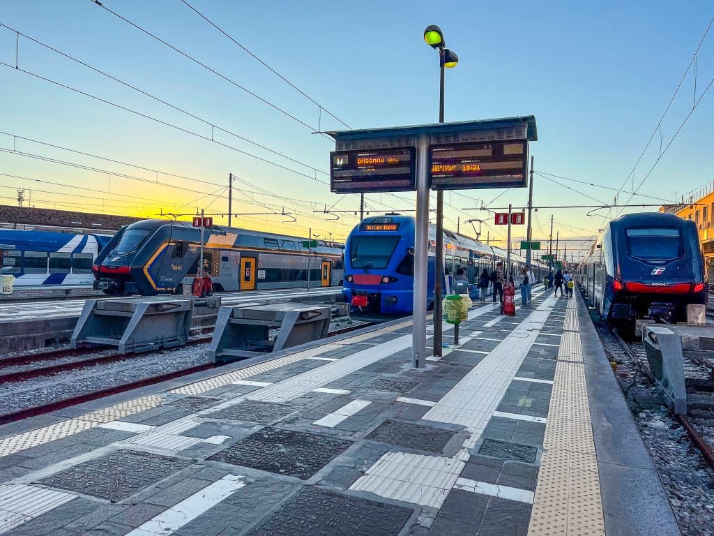 Three modern passenger trains are parked at platforms in an outdoor train station under a clear sky, with electronic signs and tactile paving—unlike Venice, where you might ask, "are there cars in Venice?" but trains like these dominate the scene.