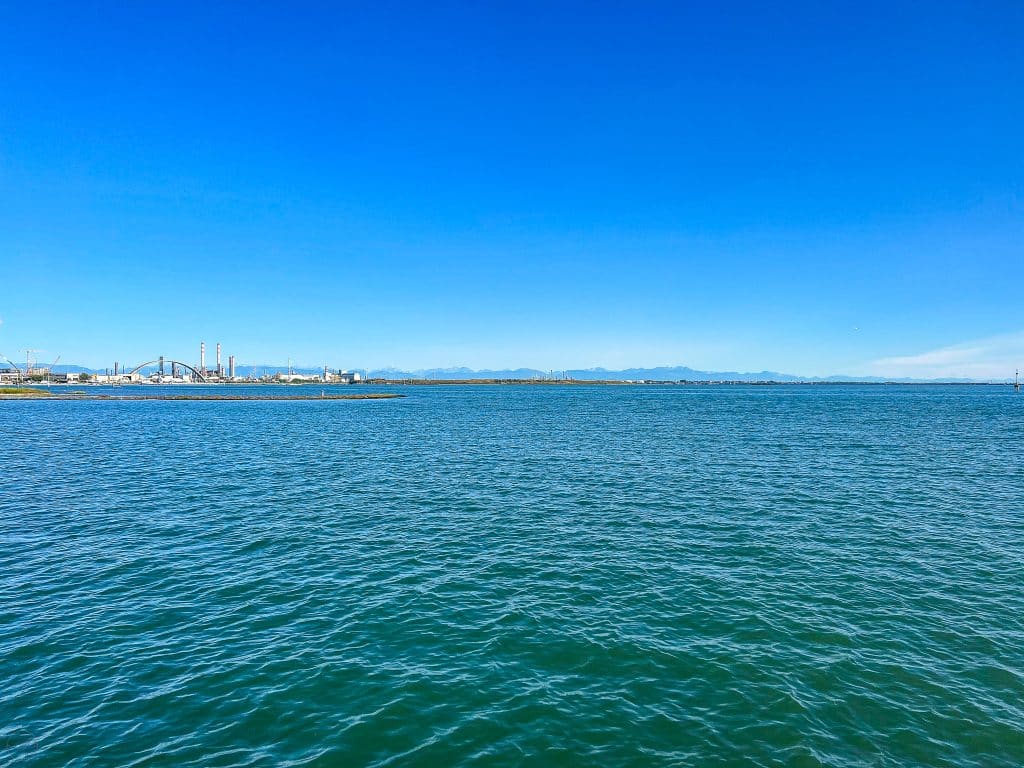 A wide expanse of blue water with an industrial facility and smokestacks visible on the distant shoreline under a clear blue sky, bringing to mind the timeless question: are there cars in Venice?.
