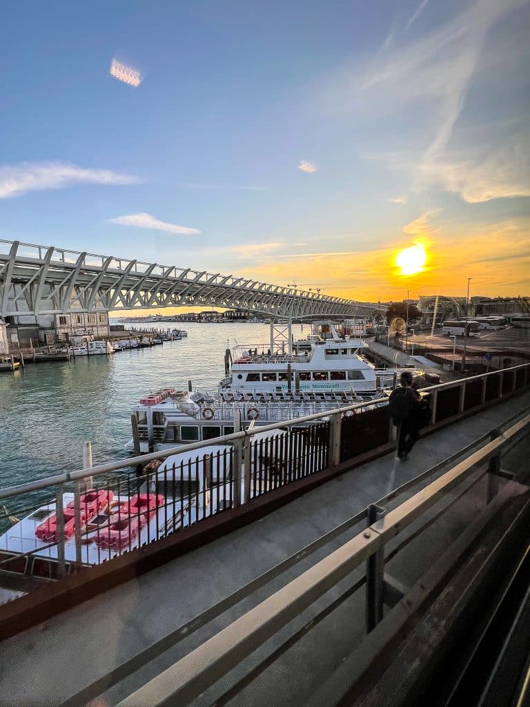 View of boats docked at a pier with a bridge overhead, water below, and the sun setting on the horizon; a person walks along the waterfront, reminding visitors that unlike cities with cars, Venice moves to the rhythm of its waterways.