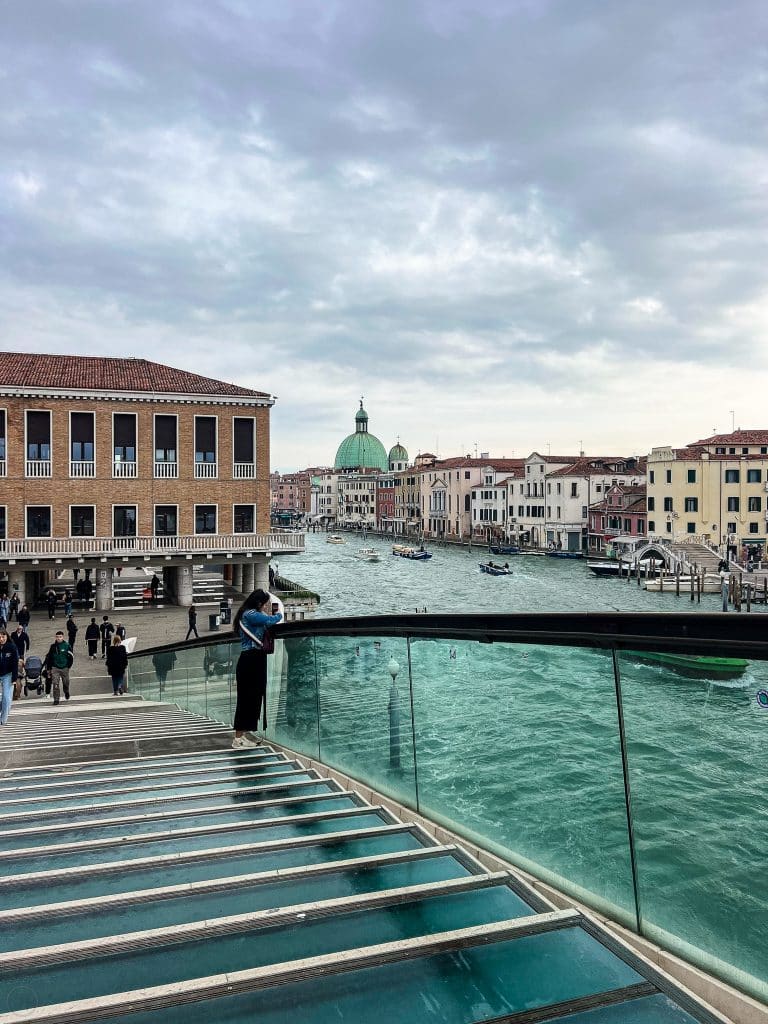 View from a glass bridge over a canal in Venice, Italy—where there are no cars—showing people walking, historic buildings, and a dome in the background under a cloudy sky.