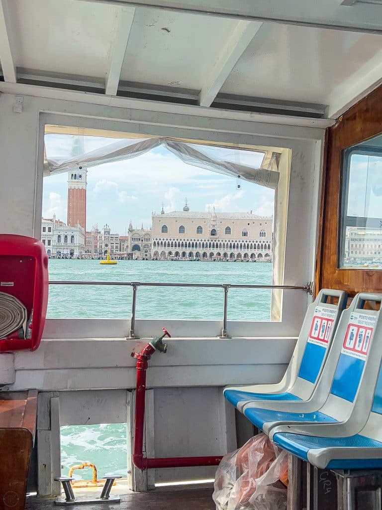 View of Venetian waterfront buildings, including St Mark’s Campanile, seen through a window from inside a boat with blue seats—reminding visitors that there are no cars in Venice, only boats gliding by emergency equipment.