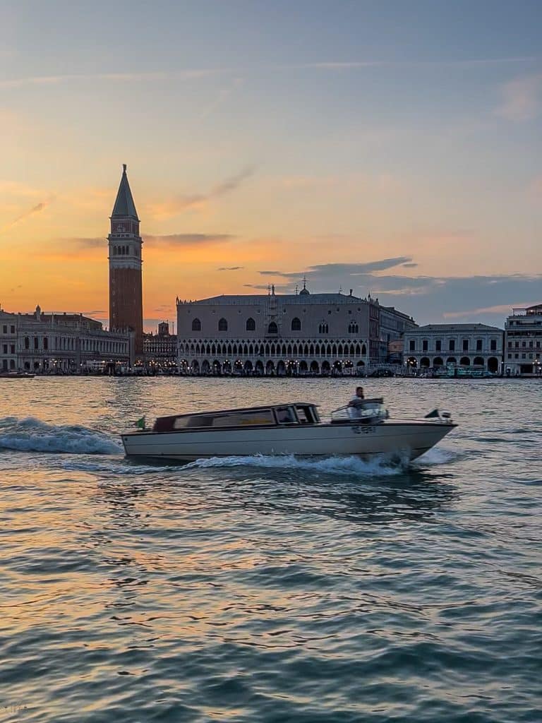 A motorboat moves across a canal in Venice at sunset, with the Campanile di San Marco and historic buildings in the background—a reminder that there are no cars in Venice, only boats gliding through its scenic waterways.