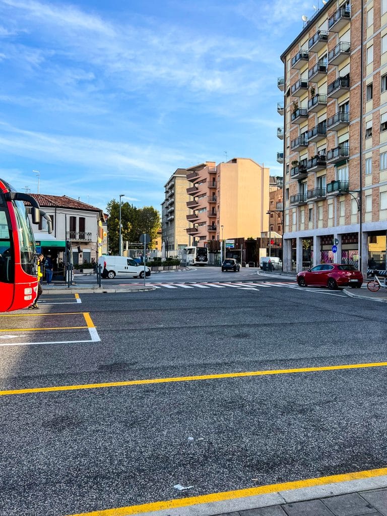 Urban intersection with apartment buildings, a red car (unlike the cars you rarely see in Venice), a red bus on the left, parked bicycles, and a few pedestrians under a clear blue sky.