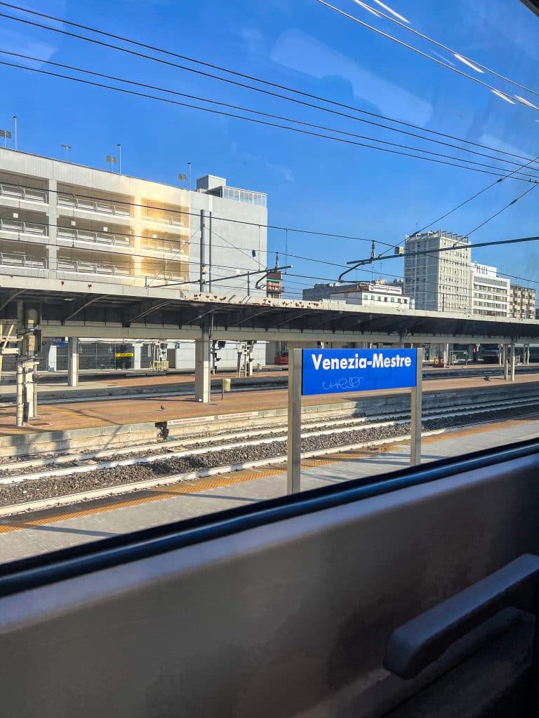 A view from a train window shows the Venezia-Mestre station sign, train tracks, and nearby buildings under a clear blue sky—a reminder that if you're wondering "are there cars in Venice," Mestre is where most visitors park before entering car-free Venice.