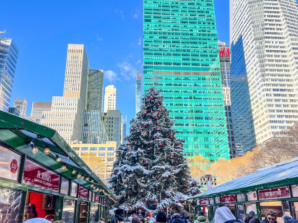 A snow-dusted Christmas tree with red ornaments stands in a city plaza near holiday market stalls, a highlight of many New York City Christmas tours.