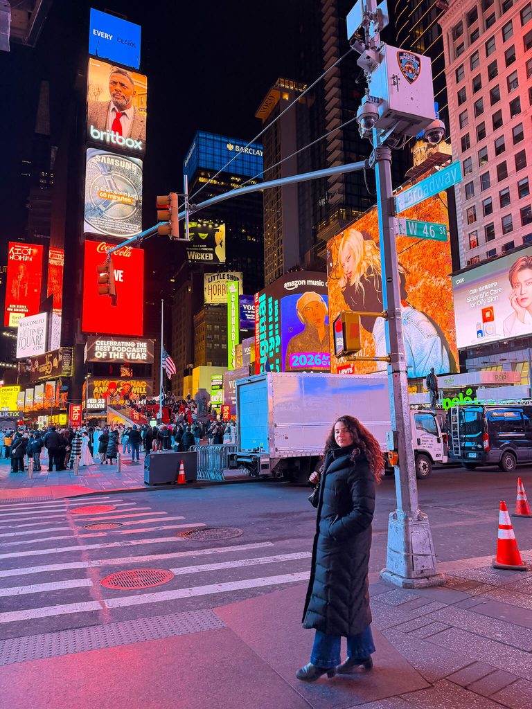 A person in a long black coat stands on a street corner in Times Square at night amidst city traffic and glowing billboards during a light snowfall in New York City.