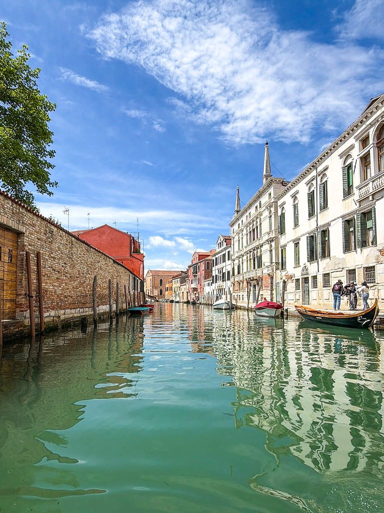 A quiet canal in Venice, where one might wonder if it smells, is lined by historic buildings and brick walls with a few small boats moored along the water.