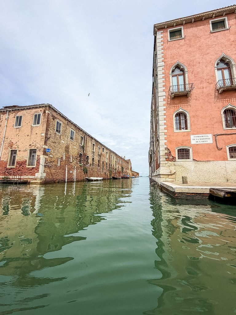 A narrow canal in Venice, where one might wonder if Venice smells, flows between a brick building and a pink one bearing a sign for “Fondamenta de la Madonna de l’Orto.”