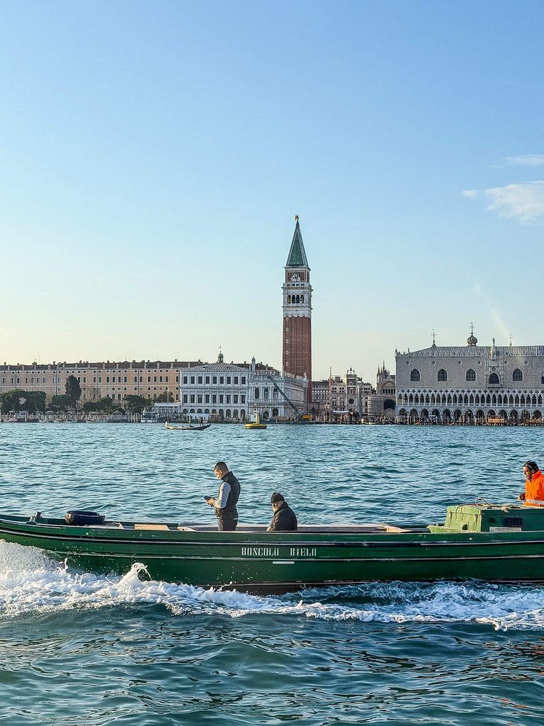 A green motorboat carries three people across the Venetian lagoon, where some wonder if Venice smells, with St. Mark’s Campanile and the Doge’s Palace in the background.
