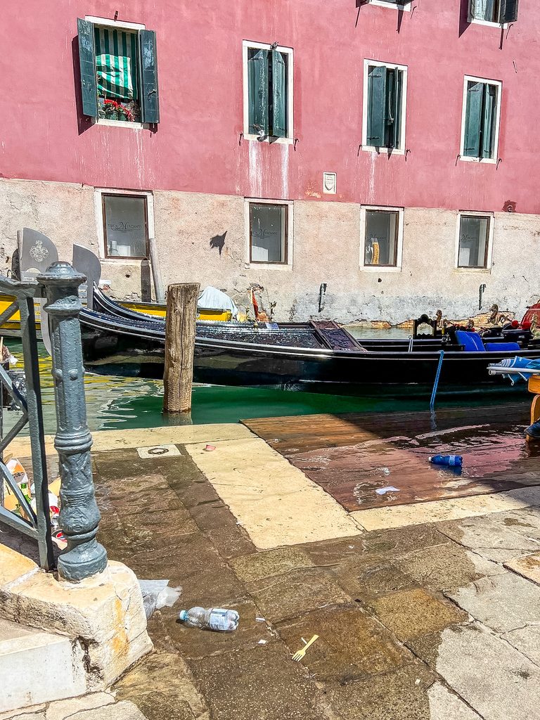 A gondola moored at a stone dock in front of a pink building in Venice, a city where visitors often wonder if the water does Venice smell.